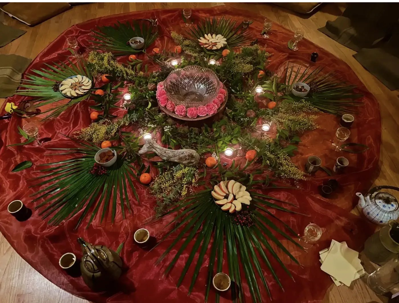 A community altar: a red cloth circle laid with palm fronds, greenery, candles, mandarins, sliced apples, a driftwood whale and a crystal bowl ringed with pink carnations